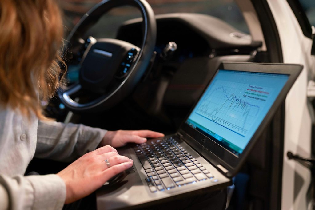 Image shows close up of a laptop balancing on a person's lap as they sit in the front seat of a vehicle with the door open. The person's face is not visible, but they appear to be female. The screen on the laptop shows a graph to suggest that an engineer can manage functions such as broadband amplifier attenuation remotely. 
