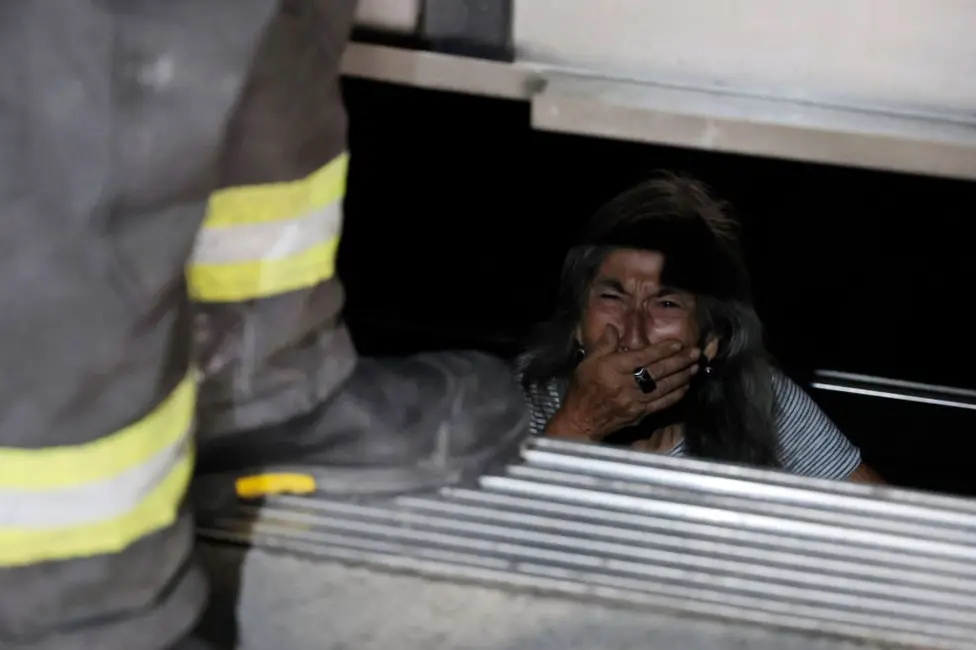 A photo inside a cramped dark area below ground level shows a woman crying and covering her mouth with her hand as she peeks out into the light. A pair of firefighter's boots and the bottom of their protective trousers in the foreground in front of her are out of focus.